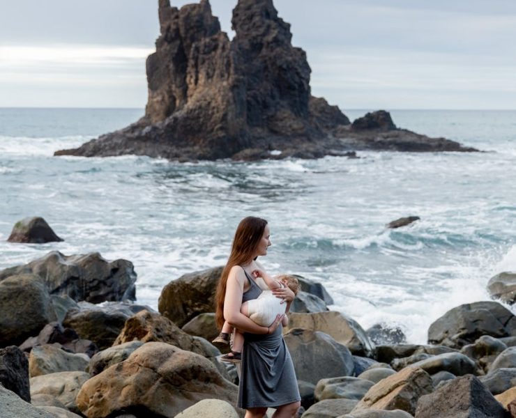 young mother with little baby on hand while standing on rocky seashore